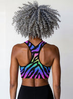 Woman showing the back of the Technicolour Essential Active Bra with bold neon green, blue, and pink zebra stripes, standing against a plain background