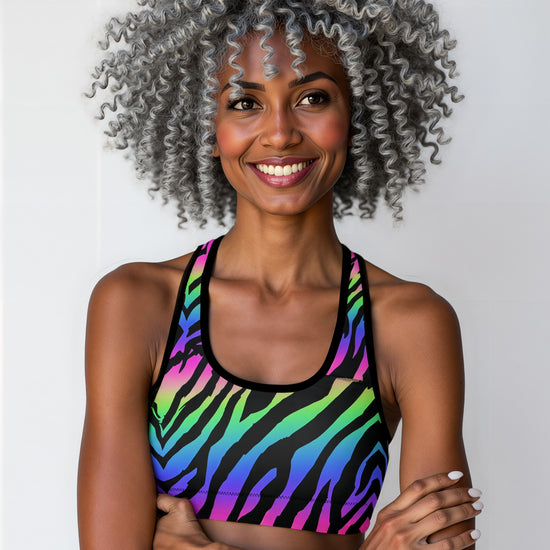 Woman wearing Technicolour Essential Active Bra with bold neon green, blue, and pink zebra stripes, smiling and standing against a plain background