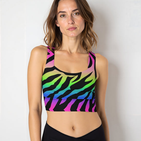 Woman wearing a Technicolour Longline Sports Bra with a bold rainbow gradient and black zebra stripe pattern, paired with high-waisted black leggings, standing against a plain light background.
