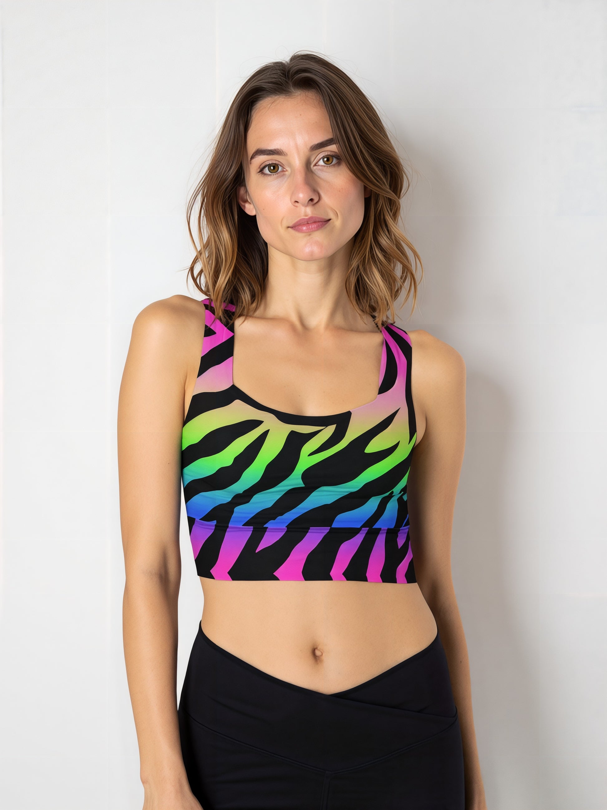 Woman wearing a Technicolour Longline Sports Bra with a bold rainbow gradient and black zebra stripe pattern, paired with high-waisted black leggings, standing against a plain light background.