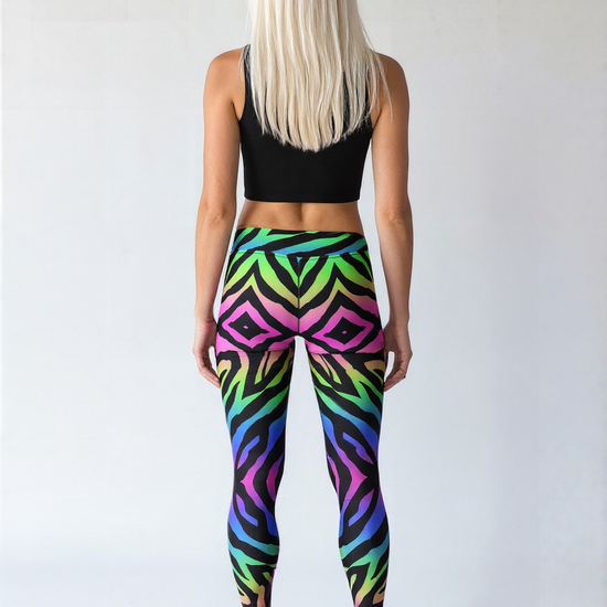 Athletic woman with long straight white hair and fair but tanned skin, shown from the back. She is wearing a black crop top, vibrant technicolour zebra-print yoga leggings, and black lace-up boots, standing against a plain light background.