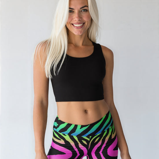 Smiling athletic woman with long straight white hair, fair but tanned skin, wearing a black crop top and vibrant technicolour zebra-print yoga leggings, standing against a plain light background.