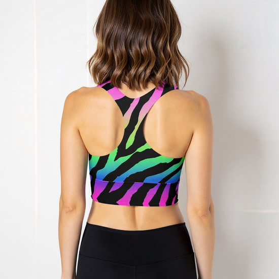 Woman wearing a Technicolour Longline Sports Bra with a bold rainbow gradient and black zebra stripe pattern, paired with high-waisted black leggings, standing against a plain light background.