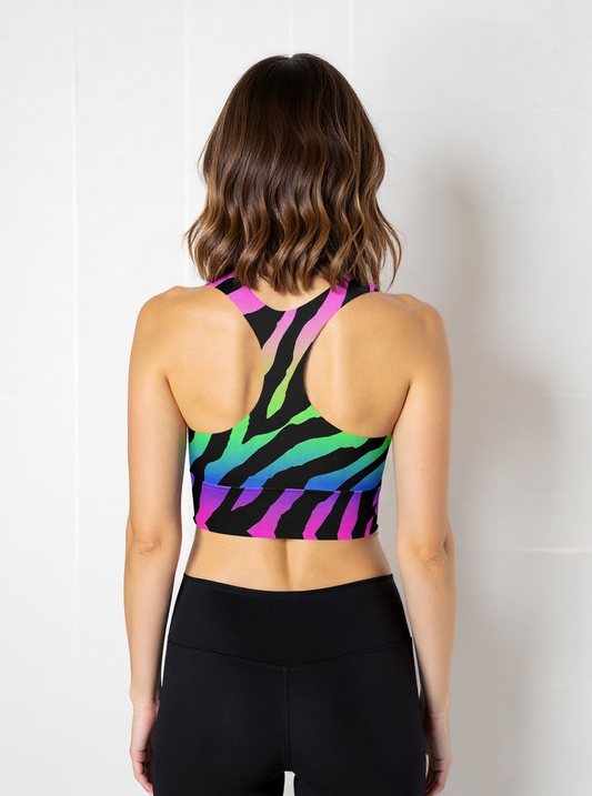 Woman wearing a Technicolour Longline Sports Bra with a bold rainbow gradient and black zebra stripe pattern, paired with high-waisted black leggings, standing against a plain light background.