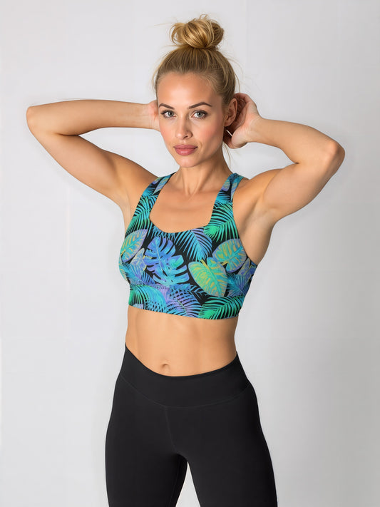 Woman wearing a Tropical Flow longline sports bra with a vibrant green and blue leaf pattern, paired with high-waisted black leggings, standing with hands behind her head against a plain background.