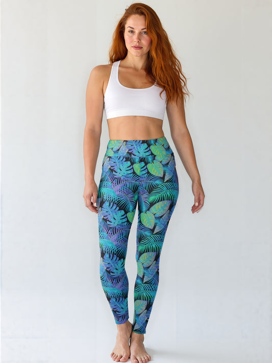 Woman wearing Tropical Flow Yoga leggings with a vibrant tropical leaf pattern in blue, green, and purple hues, paired with a white sports bra. She stands barefoot against a clean white backdrop.