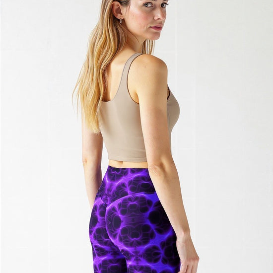 Back view of woman in high‑waisted biker shorts with a vivid purple and black abstract print, paired with a beige sleeveless crop top, standing outdoors in a relaxed, confident stance.