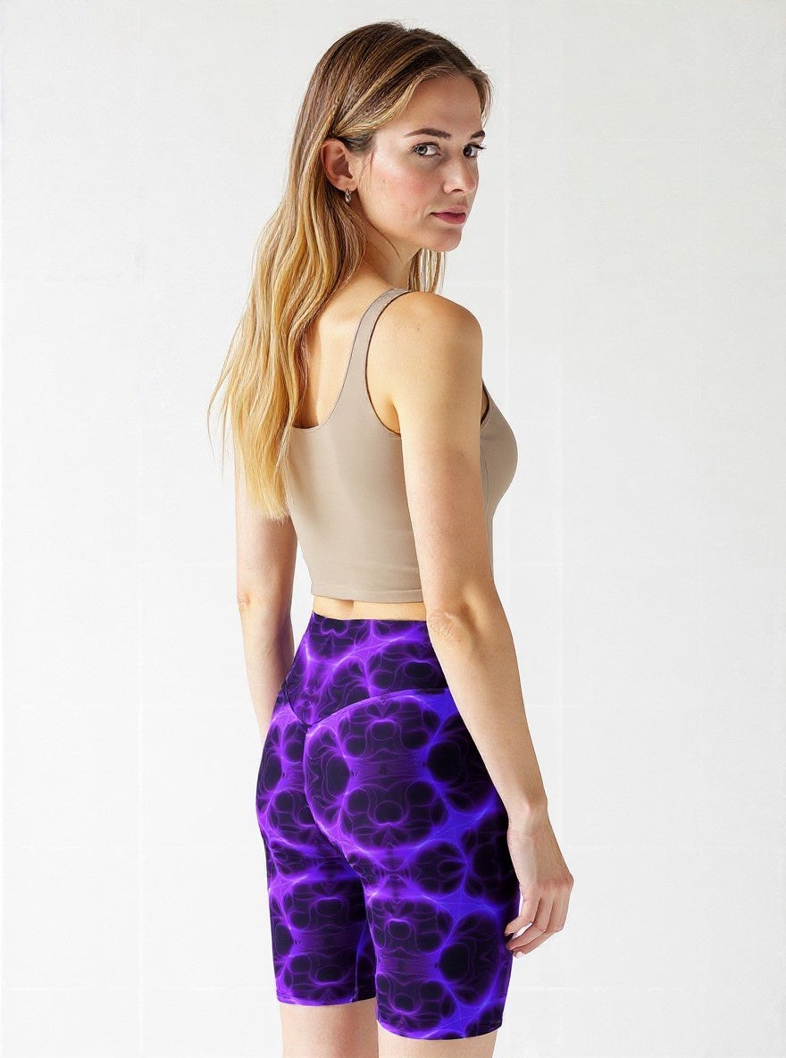 Back view of woman in high‑waisted biker shorts with a vivid purple and black abstract print, paired with a beige sleeveless crop top, standing outdoors in a relaxed, confident stance.