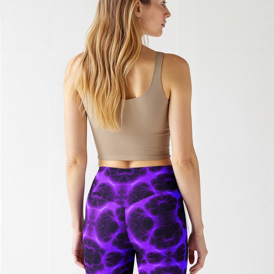 Back view of woman wearing high‑waisted biker shorts in a vivid purple and black abstract pattern, paired with a beige sports bra, standing with long wavy hair cascading over her shoulders.
