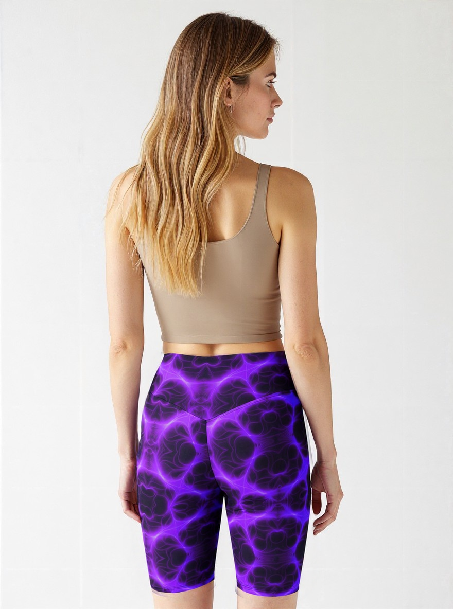 Back view of woman wearing high‑waisted biker shorts in a vivid purple and black abstract pattern, paired with a beige sports bra, standing with long wavy hair cascading over her shoulders.