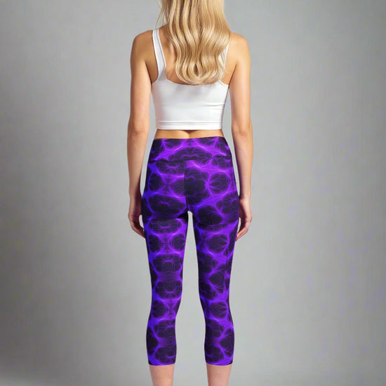 Back view of woman wearing Violet Voltage Yoga Capri leggings with a vivid purple and black lightning-inspired pattern; paired with a white crop top and black athletic shoes, standing against a light grey background.