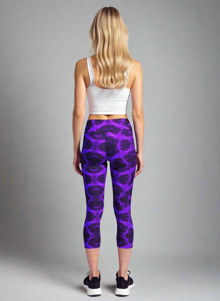 Back view of woman wearing Violet Voltage Yoga Capri leggings with a vivid purple and black lightning-inspired pattern; paired with a white crop top and black athletic shoes, standing against a light grey background.