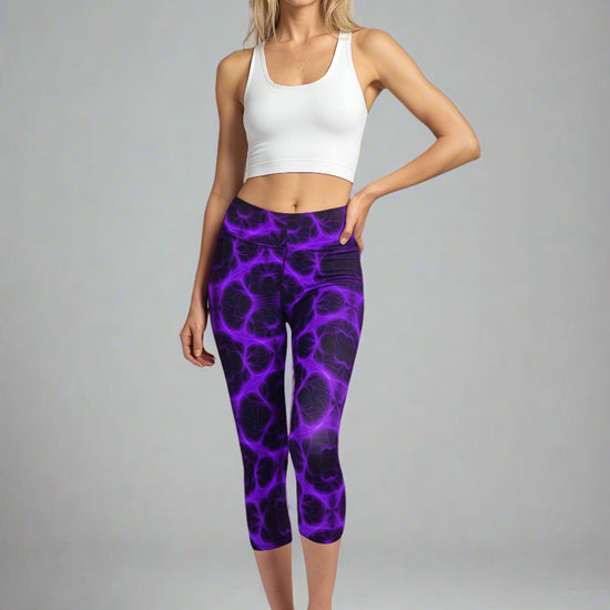Woman standing in Violet Voltage Yoga Capri leggings featuring a vivid purple and black electric swirl pattern; paired with a white crop top and black athletic shoes, set against a neutral background.