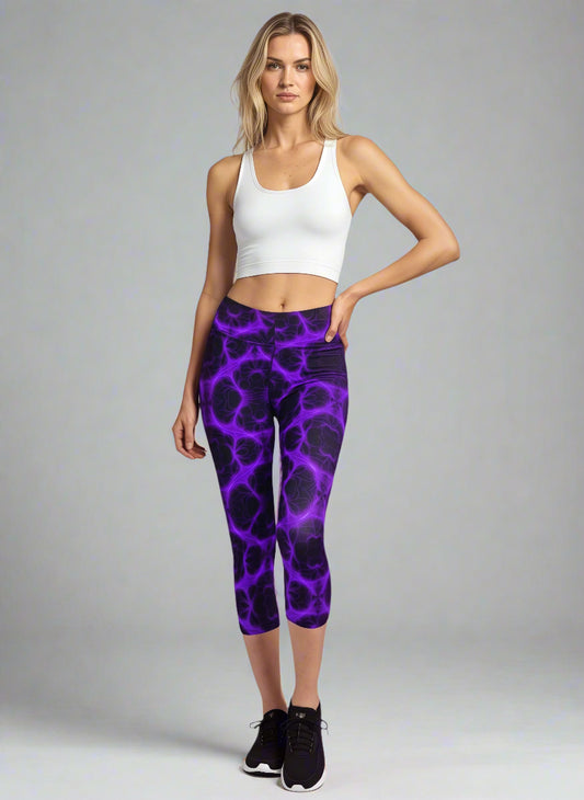 Woman standing in Violet Voltage Yoga Capri leggings featuring a vivid purple and black electric swirl pattern; paired with a white crop top and black athletic shoes, set against a neutral background.