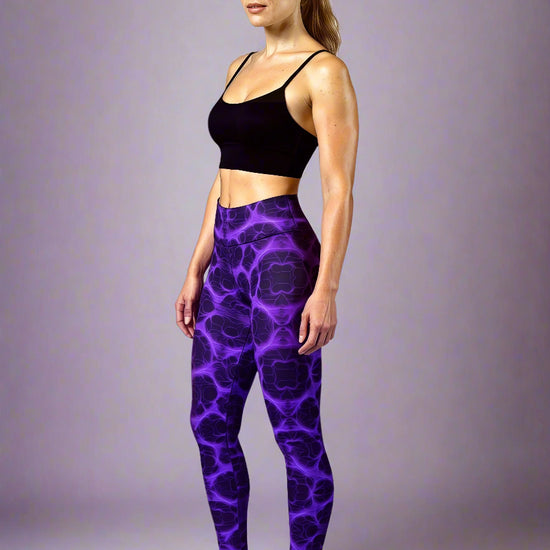 Front view of model wearing Violet Voltage Yoga Leggings, featuring a bold symmetrical pattern in electric purple and black. High‑waisted design with a glowing, abstract fractal‑inspired print, paired with a black crop top, styled barefoot against a plain light background.
