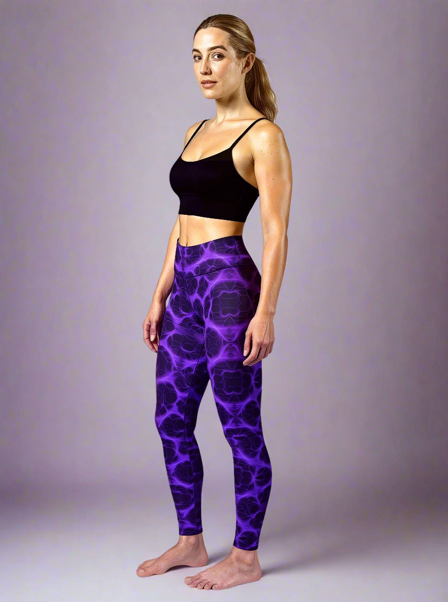 Front view of model wearing Violet Voltage Yoga Leggings, featuring a bold symmetrical pattern in electric purple and black. High‑waisted design with a glowing, abstract fractal‑inspired print, paired with a black crop top, styled barefoot against a plain light background.