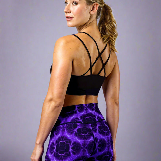 Back three‑quarter view of model wearing Violet Voltage Yoga Leggings, featuring a bold symmetrical pattern in electric purple and black. High‑waisted design with an abstract, kaleidoscopic print, paired with a black crisscross‑strap sports bra, styled against a plain light grey background.