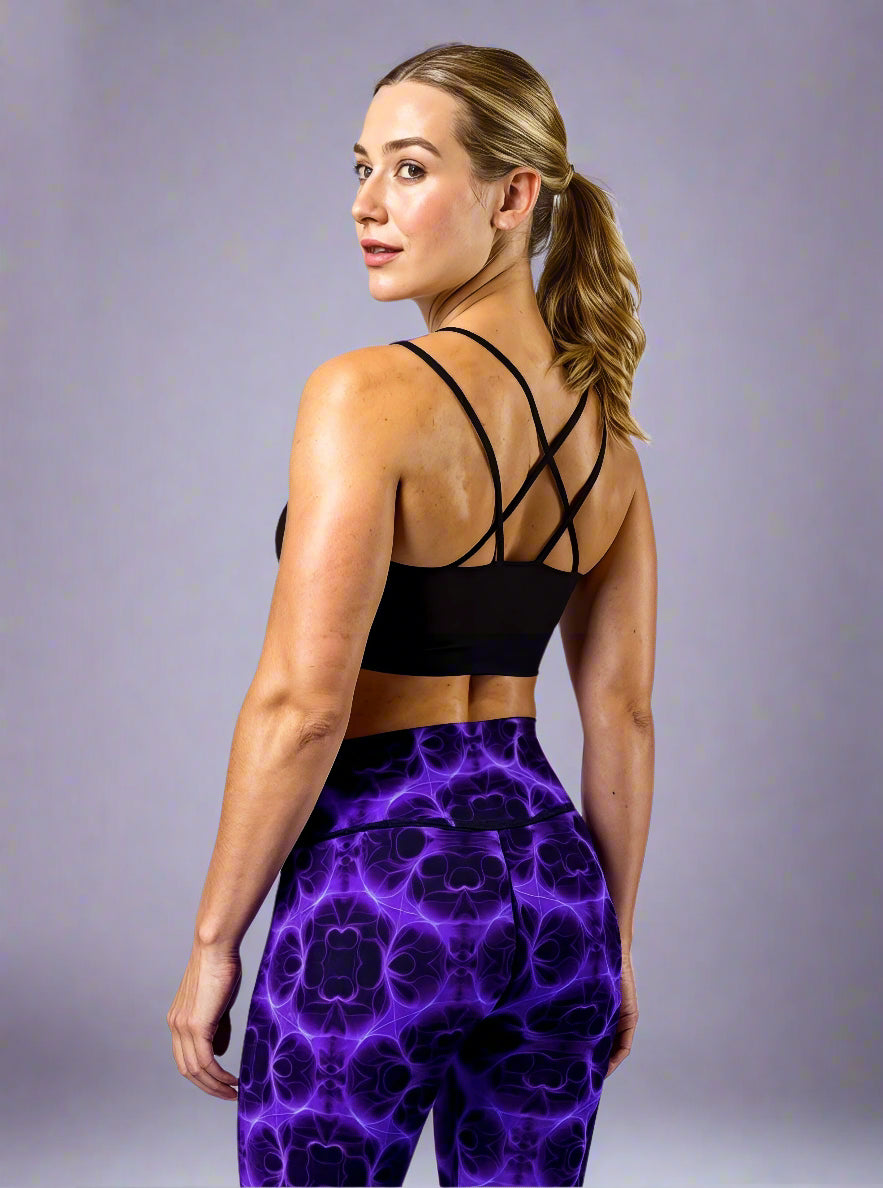 Back three‑quarter view of model wearing Violet Voltage Yoga Leggings, featuring a bold symmetrical pattern in electric purple and black. High‑waisted design with an abstract, kaleidoscopic print, paired with a black crisscross‑strap sports bra, styled against a plain light grey background.