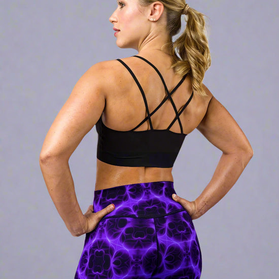 Back view of model wearing Violet Voltage Yoga Leggings, featuring a bold symmetrical pattern in electric purple and black. High‑waisted design with a glowing, abstract fractal‑inspired print, paired with a black crisscross‑strap sports bra, styled against a plain light grey background.
