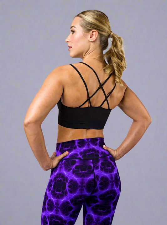 Back view of model wearing Violet Voltage Yoga Leggings, featuring a bold symmetrical pattern in electric purple and black. High‑waisted design with a glowing, abstract fractal‑inspired print, paired with a black crisscross‑strap sports bra, styled against a plain light grey background.