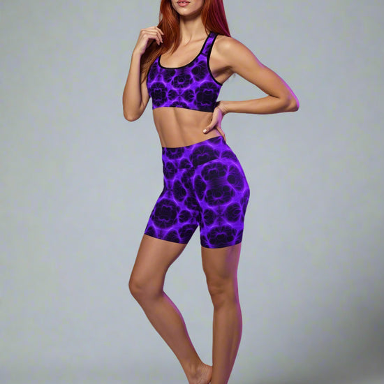 Woman wearing matching electric‑purple and black patterned sports bra and high‑waist shorts set, posing barefoot against a soft purple background — showcasing bold, luxe activewear style