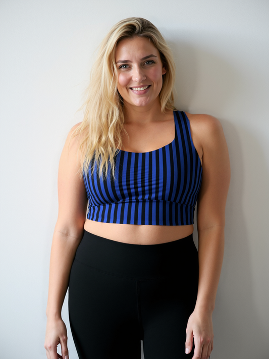 A woman with long blonde hair is smiling and standing against a white wall, wearing a Wednesday longline sports bra with blue and black vertical stripes and high-waisted black leggings.