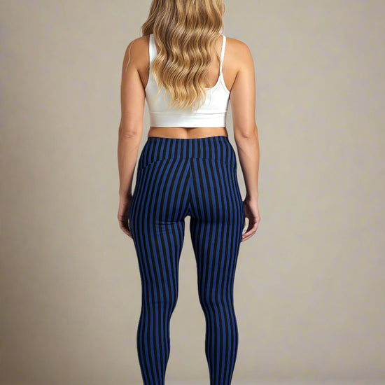 A woman with long, wavy blonde hair is shown from the back, wearing a white sports bra and blue and black vertically striped Wednesday pocket leggings, standing against a plain background with white sneakers.
