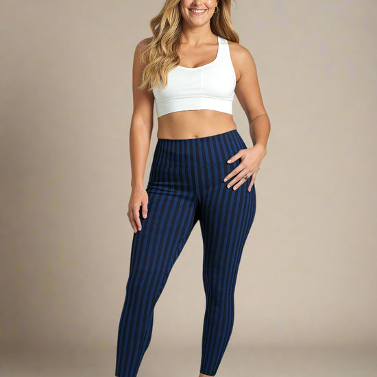 A woman with long, wavy blonde hair is smiling and standing against a plain background, wearing a white sports bra, blue and black vertically striped Wednesday pocket leggings, and white sneakers. One hand rests on her hip, showing the fit and style of the leggings.