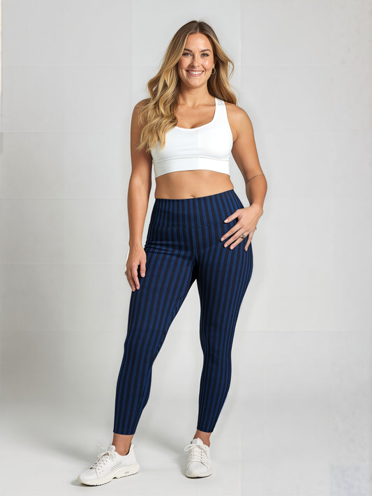 A woman with long, wavy blonde hair is smiling and standing against a plain background, wearing a white sports bra, blue and black vertically striped Wednesday pocket leggings, and white sneakers. One hand rests on her hip, showing the fit and style of the leggings.