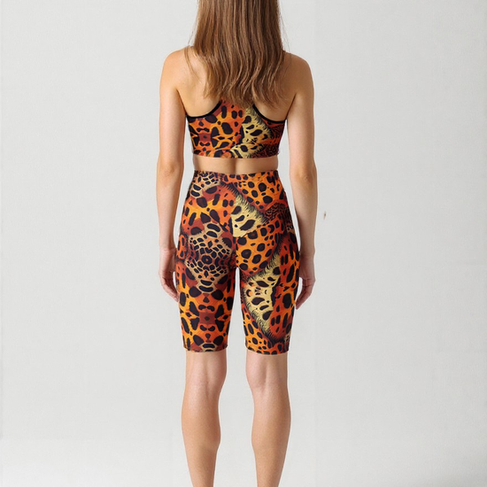 A woman is shown from behind, wearing a matching sports bra and Wildfire biker shorts set. The biker shorts feature a bold, all-over animal print pattern in orange, yellow, and black, with a high-waisted, fitted design that falls just above the knee. She is also wearing black athletic shoes, and the plain background highlights the vibrant, eye-catching details of the activewear.