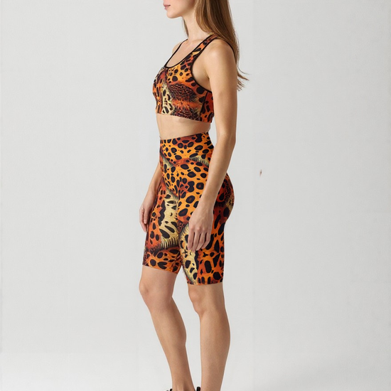 A woman is shown in profile against a plain background, wearing a matching sports bra and Wildfire biker shorts set. The high-waisted biker shorts feature a vibrant animal print pattern in orange, yellow, and black, hitting just above the knee. She pairs the outfit with black athletic sneakers, showcasing a dynamic and stylish activewear look.