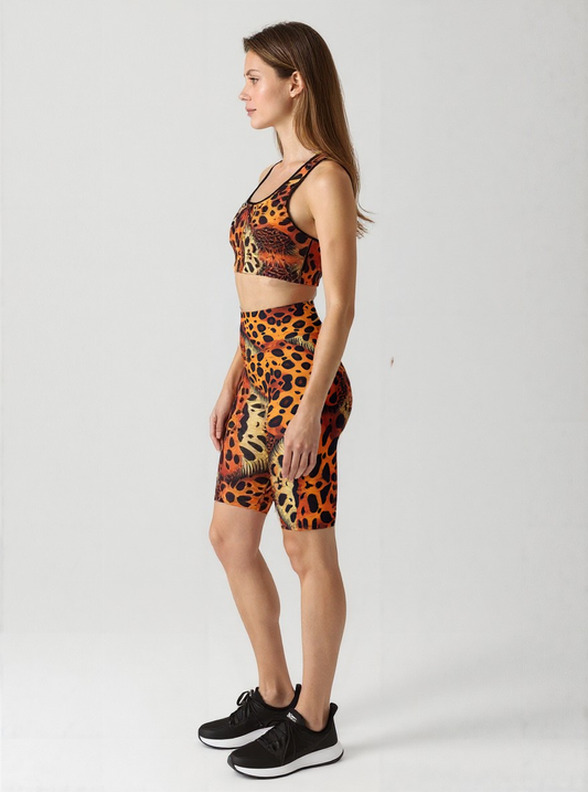 A woman is shown in profile against a plain background, wearing a matching sports bra and Wildfire biker shorts set. The high-waisted biker shorts feature a vibrant animal print pattern in orange, yellow, and black, hitting just above the knee. She pairs the outfit with black athletic sneakers, showcasing a dynamic and stylish activewear look.