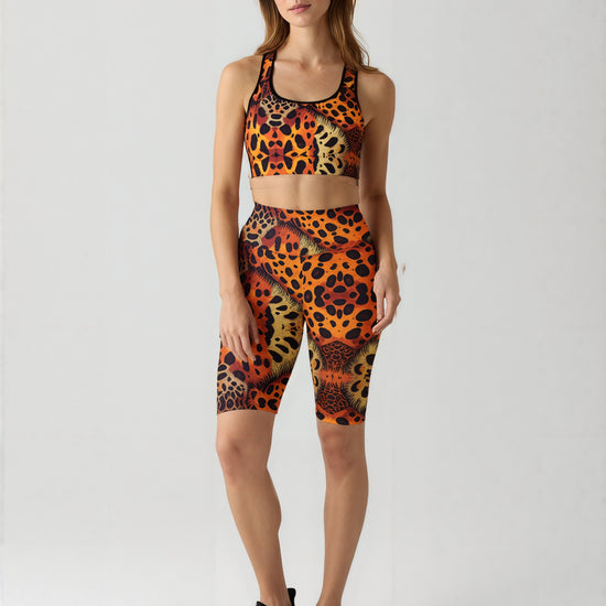 A woman stands against a plain background wearing a matching sports bra and Wildfire biker shorts, both featuring a bold animal print pattern in orange, yellow, and black. The high-waisted biker shorts have a vibrant, eye-catching design and hit just above the knee. She pairs the set with black athletic sneakers, highlighting a striking and stylish activewear look.