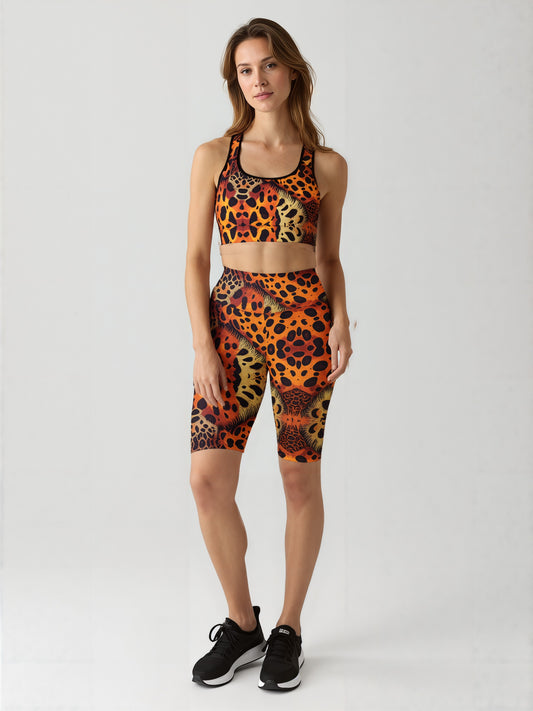 A woman stands against a plain background wearing a matching sports bra and Wildfire biker shorts, both featuring a bold animal print pattern in orange, yellow, and black. The high-waisted biker shorts have a vibrant, eye-catching design and hit just above the knee. She pairs the set with black athletic sneakers, highlighting a striking and stylish activewear look.