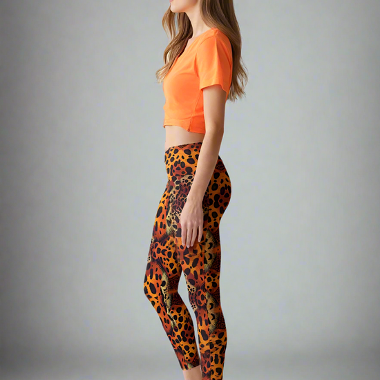 A woman is standing in profile, wearing a bright orange cropped t-shirt and Wildfire Crossover Leggings. The leggings feature a bold, vibrant animal print in shades of orange, yellow, and black, with a flattering high-rise fit. She pairs the outfit with white sneakers, and the background is plain and softly lit, emphasizing the striking leggings.